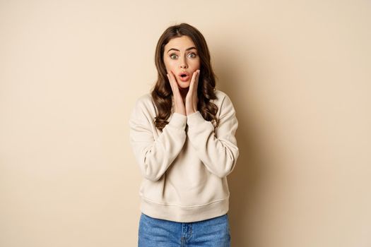 Image of surprised, amazed brunette girl gasping, looking fascinated at camera, standing over beige background.