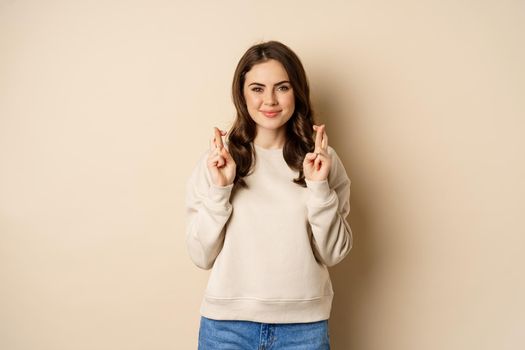Confident woman praying, making wish with fingers crossed for good luck, standing over beige background.