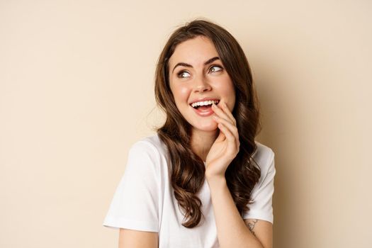 Close up portrait of coquettish beautiful girl gazing silly, gently smiling and giggling, posing flirty and cute against beige background. Women beauty and valentines concept.