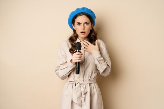 Image of insecure young woman standing with mic microphone and looking shocked worried, fear of speaking in public, standing over beige background.