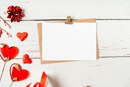 Flat lay of paper envelope with blank mockup greeting paper card. White wooden table background with Valentine day gift, letter, heart shape, lollipops, confetti, decor. Top view, mock up invitations