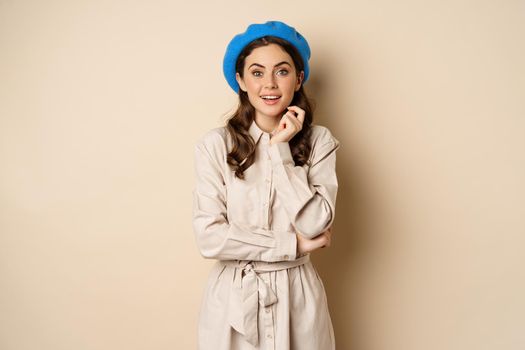 Image of stylsh beautiful woman looking surprised, shocked reaction at camera, posing in trench coat against beige background.