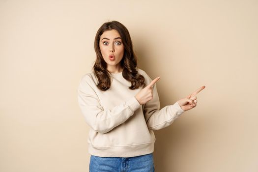 Surprised brunette woman pointing fingers right, showing advertisement and say wow impressed, standing over beige background.