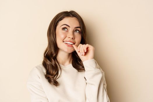 Cute modern woman thinking, daydreaming and smiling, looking up thoughtful, standing over beige background.