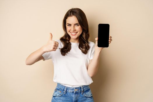 Image of happy young woman showing thumbs up and mobile phone screen, recommending webstore, online shopping application, standing pleased over beige background.
