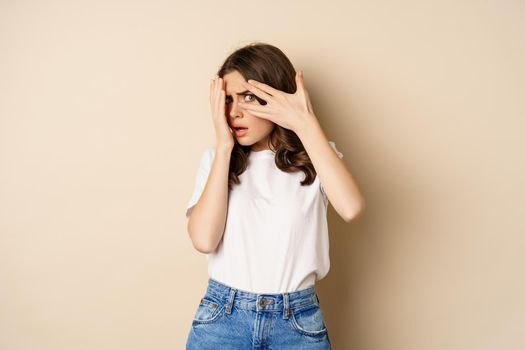 Young woman scared to watch, cover eyes with hands and peeking through fingers at scary moment, standing over beige background.