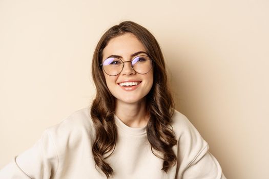 Close up portrait of beautiful modern woman in glasses, smiling and looking happy, posing in eyewear against beige background.