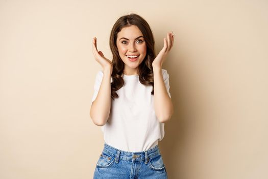 Surprise. Happy young woman open eyes with hands and waiting for gift, standing over beige background. Copy space