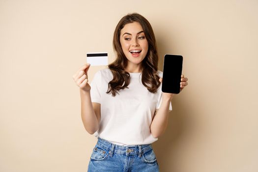 Online shopping and people concept. Young beautiful woman looking happy, showing credit card discount and mobile phone screen, standing over beige background.