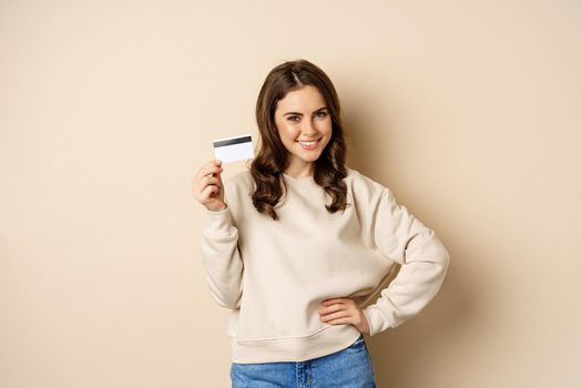 Smiling beautiful woman showing credit card, concept of shopping, contactless payment, standing over beige background.