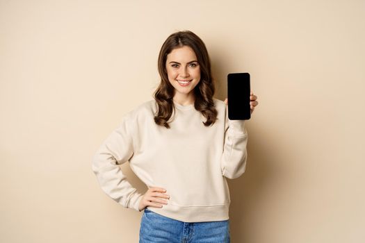 Stylish smiling woman showing smartphone screen, mobile app interface, standing over beige background.