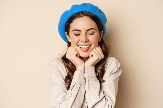 Close up portrait of feminine young woman in trendy french hat, posing cute and romantic, making silly coquettish face and tender smile, beige background.
