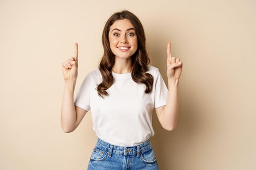 Enthusiastic young woman, female customer pointing fingers up and smiling, showing banner or logo, standing against beige background.