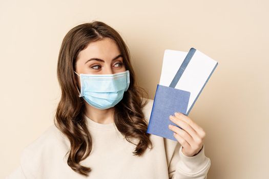 Travel and covid-19 pandemic. Close up portrait of smiling woman in medical mask, showing passport with two tickets, concept of tourism, beige background.