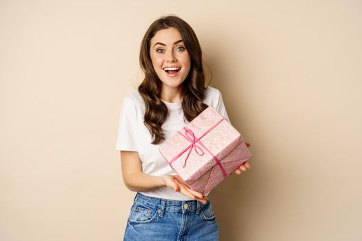 Celebration and holidays concept. Happy young woman holding gift wrapped in pink box, receive present, looking amazed and surprised, standing over beige background.