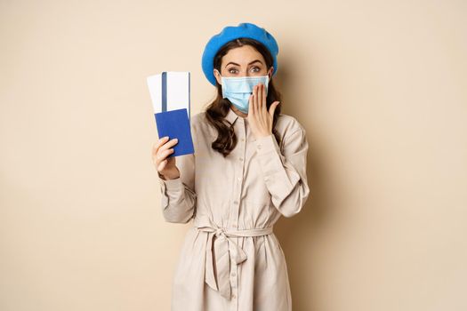 Covid pandemic and travelling concept. Portrait of cute girl in medical face mask going on trip, showing passport with tickets abroad and looking excited, beige background.