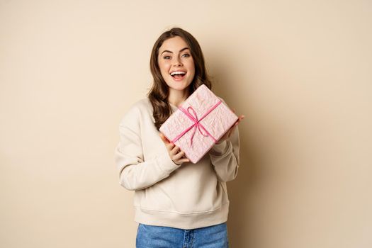 Happy beautiful girlfriend holding pink gift box and laughing, smiling joyful, concept of holidays and celebration, beige background.
