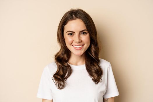People. Close up portrait of young woman smiling, looking happy, wearing casual white t-shirt, standing healthy and cheerful against beige background.