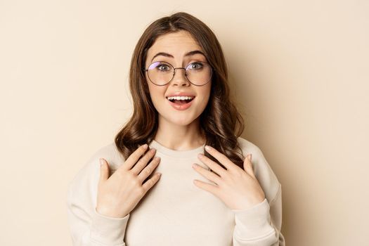 Close up portrait of beautiful girl in glasses, looking amazed and surprised, gasping excited, standing over beige background.