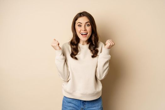 Enthusiastic brunette woman laughing and smiling, cheering, shaking hands and celebrating, triumphing, standing over beige background.