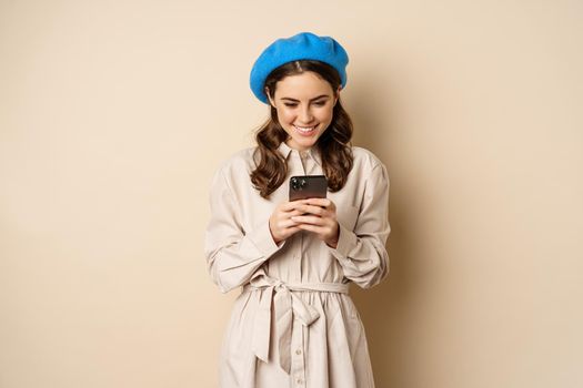 Modern girl using smartphone, smiling and posing happy with mobile phone, standing over beige background.