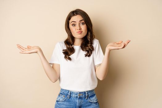 Young confused woman shrugging shoulders and with clueless face expression, standing over beige background.