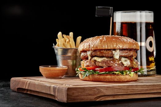 Big burger with two veal patties, creamy blue cheese, caramelized onions, fresh tomatoes and arugula in wheat sesame bun served with glass of foamy amber lager, crispy fries and spicy sauce