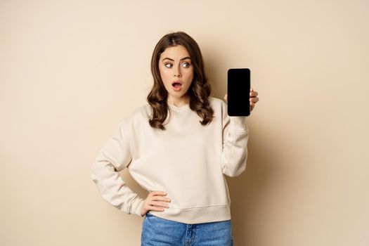 Woman showing smartphone screen and looking surprised at mobile phone, standing in sweater over beige background.
