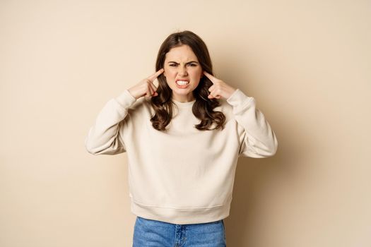 Annoyed and angry woman shut ears, complaining at loud noisy music noise, standing over beige background.