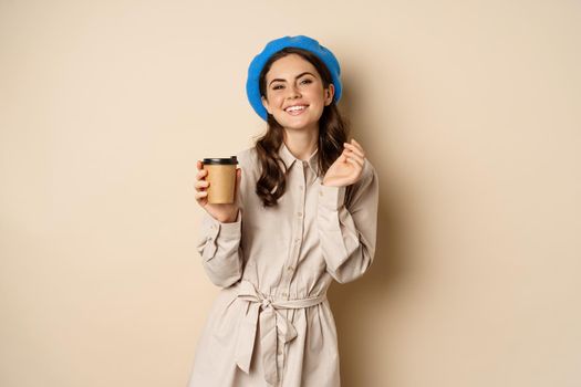 Beautiful happy girl drinking takeaway coffee from cafe and smiling, posing with cup of beverage, beige background.
