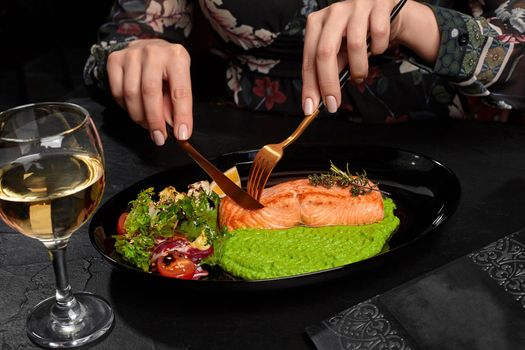 Female hands cutting appetizing grilled salmon steak served on plate with green pea puree, fresh herbs and vegetables salad, lemon slice and glass of white wine on black background