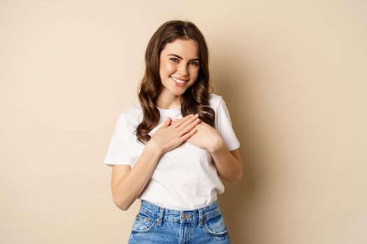 Portrait of young caucasian woman hold hands on heart and smiling, daydreaming, thinking of smth heartwarming, posing against beige background.