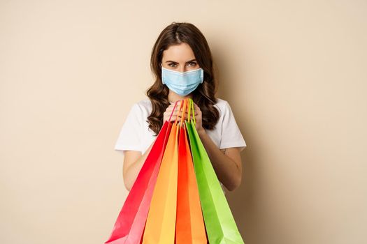 Covid-19 pandemic and lifestyle concept. Young woman posing in medical face mask with shopping bags from mall, vaccinated girl going in store in personal protective equipment, beige background.