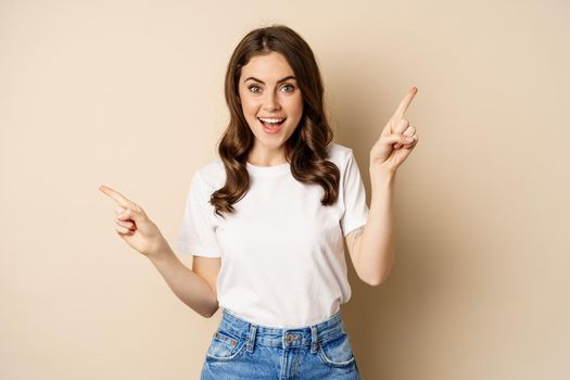 Happy authentic girl smiling, pointing fingers sideways, showing left and right banner, demonstrating promo, standing against beige background.