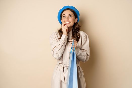 Image of modern beautiful woman posig with shopping gift bag, smiling and looking up thoughtful, thinking, standing over beige background.