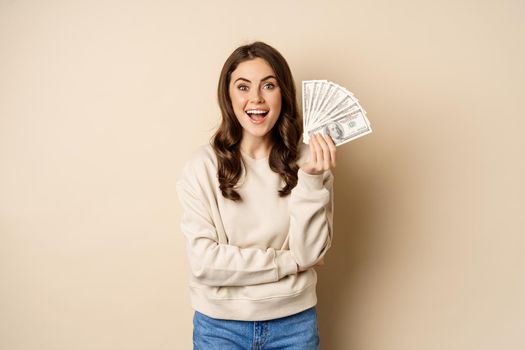 Happy woman showing money, cash and smiling pleased, concept of microcredit and shopping, standing over beige background.