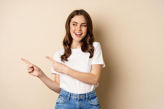 Stylish young caucasian woman smiling, pointing fingers left, showing advertisement, promo offer, standing against beige background.