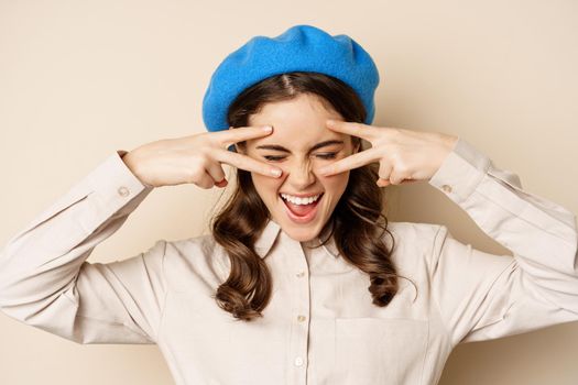 Close up portrait of carefree happy woman, showing peace v-sign and laughing, having fun, standing against beige background.