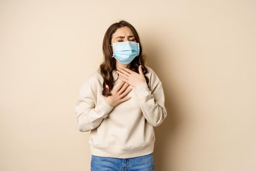 Covid-19 and health concept. Sick woman in medical face mask coughing, feeling ill with sour throat, standing over beige background.