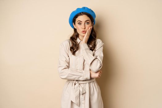 Image of stylsh beautiful woman looking surprised, shocked reaction at camera, posing in trench coat against beige background.