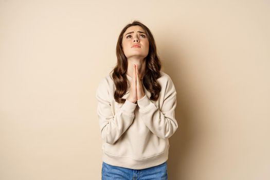 Woman begging and pleading god, supplicating, say please, standing desperate over beige background. Copy space