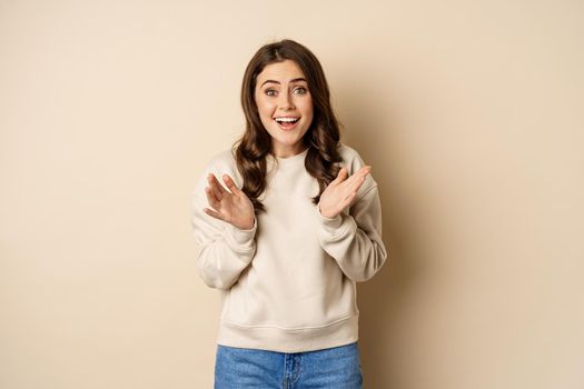 Image of surprised, amazed brunette girl gasping, looking fascinated at camera, standing over beige background.