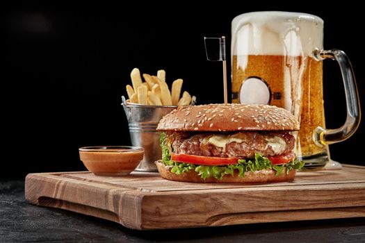 Tasty burger with juicy beef patty, creamy blue cheese, caramelized onion, fresh vegetables in fluffy bun with sesame served with glass of pale lager, crispy french fries and sauce on wooden board