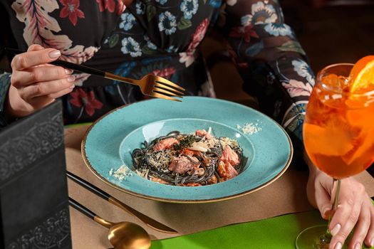 Female hands holding glass of orange spritz cocktail and fork going to eat delicious squid ink pasta with seafood and grated parmesan during dinner in restaurant, cropped image