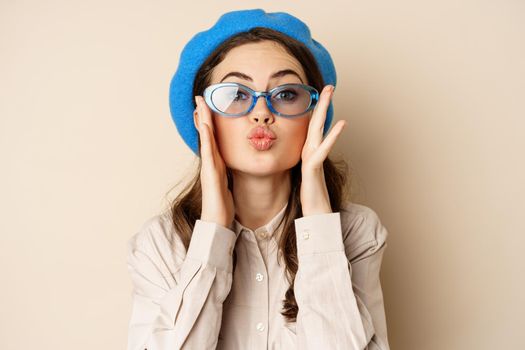 Beautiful stylish woman in sunglasses, blowing air kiss on hands near lips, gazing softly and flirty at camera, posing against beige background.