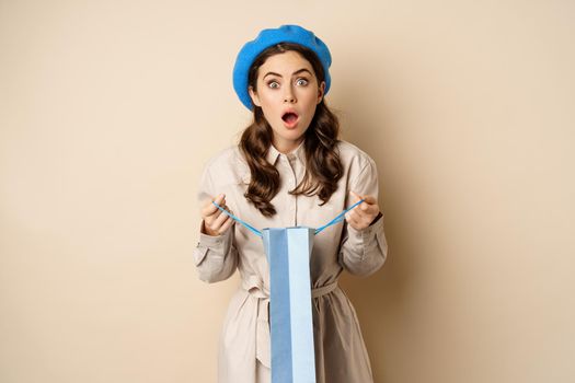 Image of girl looking surprised after opening gift bag, looking at present amazed, standing in stylish outfit over beige background.