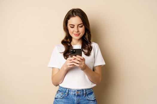 Young modern girl chatting on app, using smartphone app and smiling, standing over beige background.