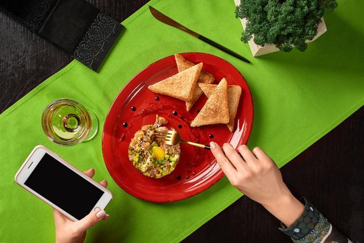 Cropped image of woman hands holding mobile phone and fork going to eat delicious salmon tartare with avocado, capers, gherkins, soy sauce and quail yolk served with crispy toasts and glass of wine