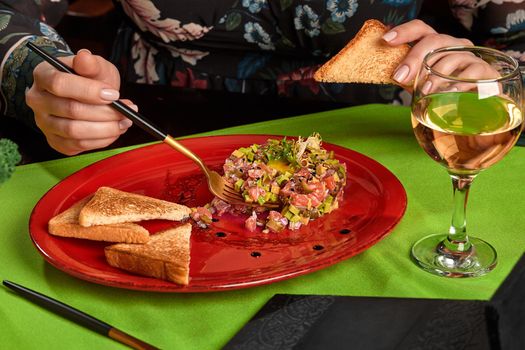 Female hands holding crispy toasted bread and fork, taking salmon tartare from plate during dinner with wine in restaurant, cropped shot