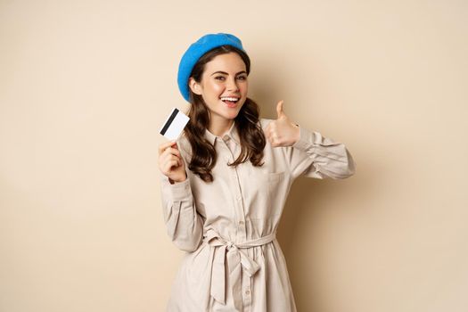 Concept of shopping and sale. Happy young woman in stylish clothes, showing credit card store discount and thumbs up, recommending, standing over beige background.